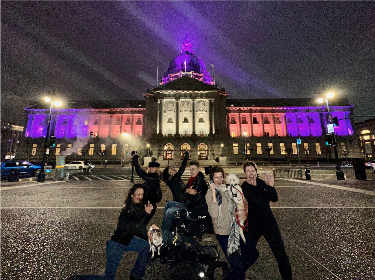 Women standing in front of San Francisco's City Hall building which is lit in the colors of the lesbian flag